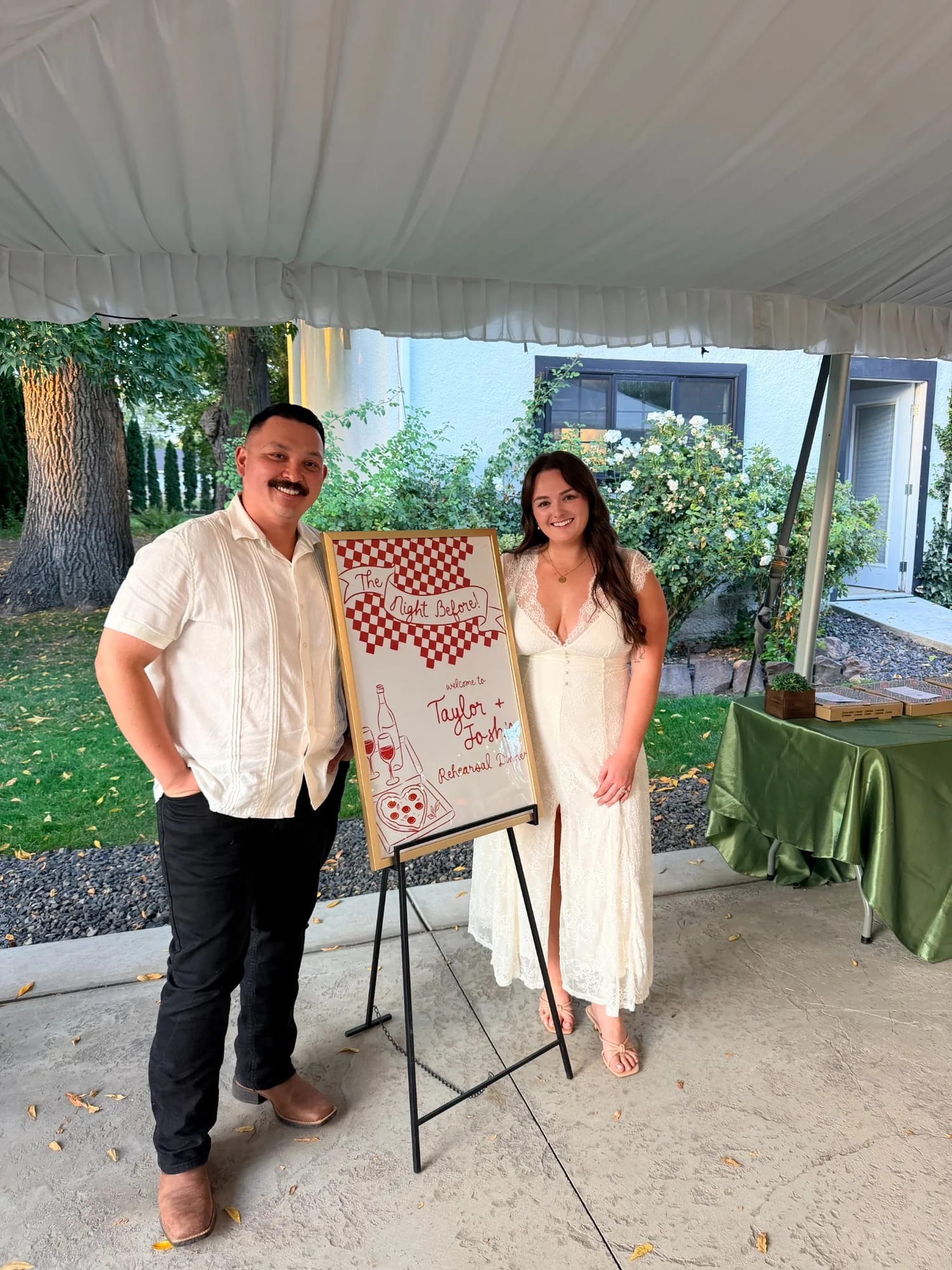 A couple beside their rehearsal dinner welcome sign and Margherita Pizza catering table